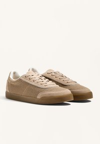 Beige színű sportcipő hálós anyaggal, velúr részletekkel, lapos fűzőkkel és gumitalppal. Jellemzője a világos színű varrás és az alacsony profilú dizájn.