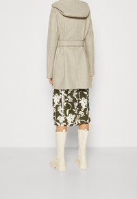 Cappotto di lana beige con cappuccio, cinturato in vita, abbinato a una gonna floreale verde scuro e stivali bianchi fino al ginocchio.