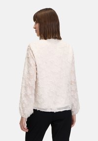 Hellrosa Bluse mit einem strukturierten Blumenmuster, transparentem Material, Ballonärmeln und geradem Schnitt am Saum. Kombiniert mit schwarzen Hosen.