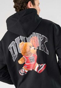 Zwarte hoodie met een grote afbeelding van een beer in rode basketbalkleding, met het woord "DUNK" erboven. Zachte stof, losse pasvorm.