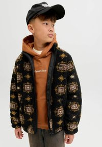 Flis jakna s crnom bazom, uzorkovana zemljanim tonovima i geometrijskim oblicima, složena preko narančaste dukserice, uparen s crnom kapom.