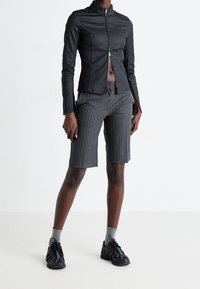Sort lynlåsjakke i tætsiddende stil med høj krave, parret med grå stribede shorts. Sorte træningssko og grå sokker fuldender outfittet.
