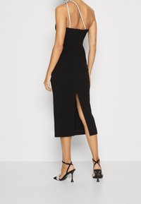 Černé midi šaty s elegantním střihem, s rozparkem na boku a zdobenými ramínky na tenkých průvlecích. Dokončení outfitu tvoří černé lodičky s ozdobnými detaily.