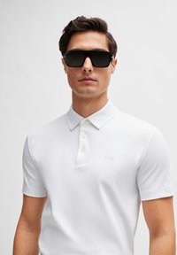 White short-sleeve polo shirt with a straight collar, three-button placket, and embossed logo on the chest; model wearing black sunglasses.