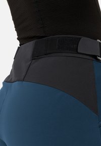 Blauwe technische broek met zwarte accenten, voorzien van een textuur in het taillegebied en klittenbandsluitingen, ontworpen voor mobiliteit en comfort.