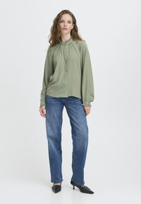 Woman wearing a light green ruffled collar blouse, blue straight-leg jeans, and black pointed-toe heels standing against a plain white background.