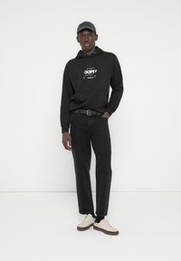 Νέος άνδρας που φοράει μαύρη μπλούζα με κουκούλα και λογότυπο, μαύρο παντελόνι, μαύρο καπέλο και λευκά αθλητικά παπούτσια, στέκεται μπροστά σε λευκό φόντο.