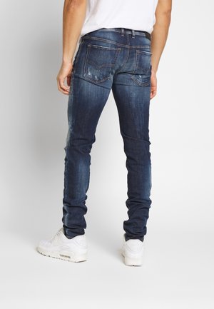 Mand iført mørkeblå slim-fit jeans med falmede og slidte detaljer, hvide sneakers og en hvid t-shirt, der står mod en ensfarvet baggrund.
