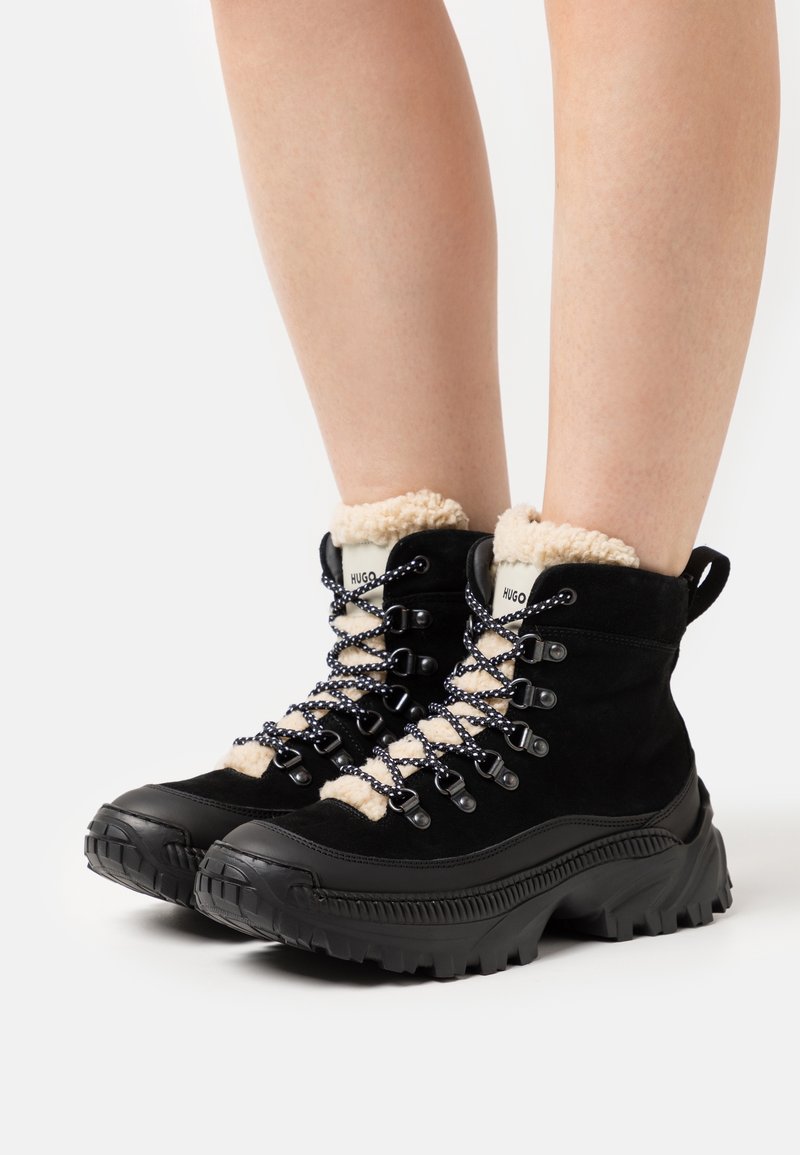 Bottes d'hiver noires à lacets avec semelles épaisses et doublure en polaire beige, portées aux pieds sur fond blanc.