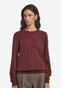 Burgunderfarbener Pullover mit rundem Ausschnitt, gerippten Bündchen und Saum; aus weichem, strukturiertem Material; kombiniert mit hellgrauen, plissierten Hosen.