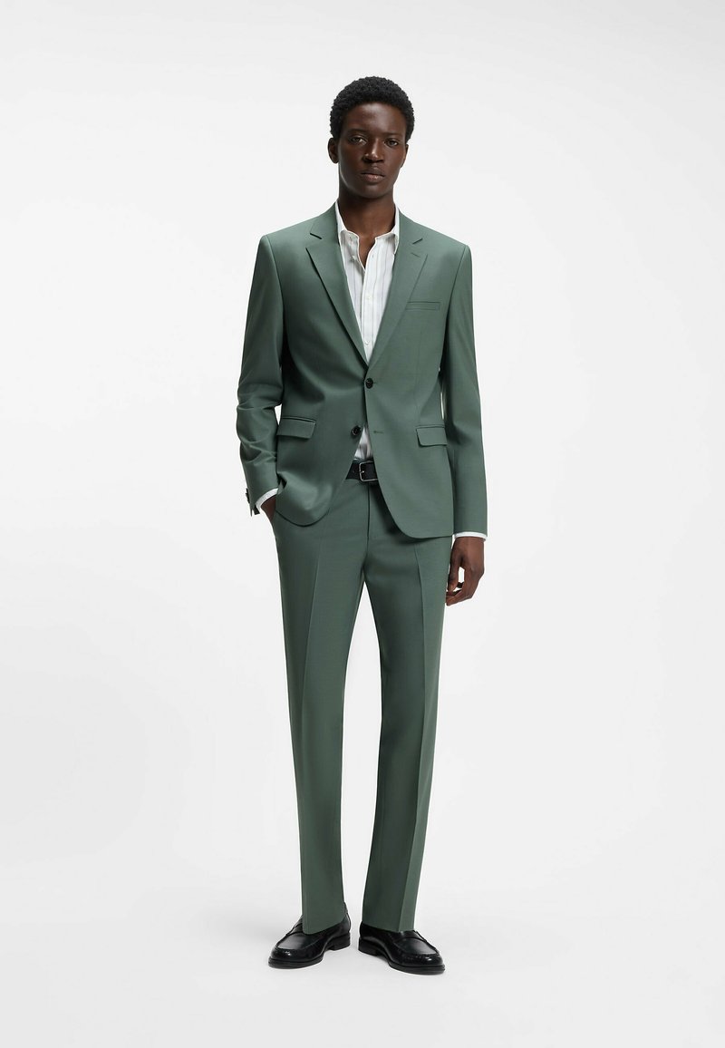 Jeune homme debout les mains dans les poches, portant un costume vert, une chemise blanche rayée, une ceinture noire et des chaussures habillées noires sur fond blanc.
