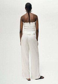 Top bianco senza spalline con dettagli texturizzati e orlo arricciato, abbinato a pantaloni bianchi larghi e sandali neri. Capelli in una lunga treccia.