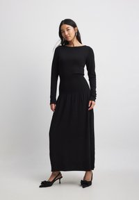 Black long-sleeve dress with a fitted top and gathered waist, flowing down to a straight skirt. Smooth fabric with minimal detailing.
