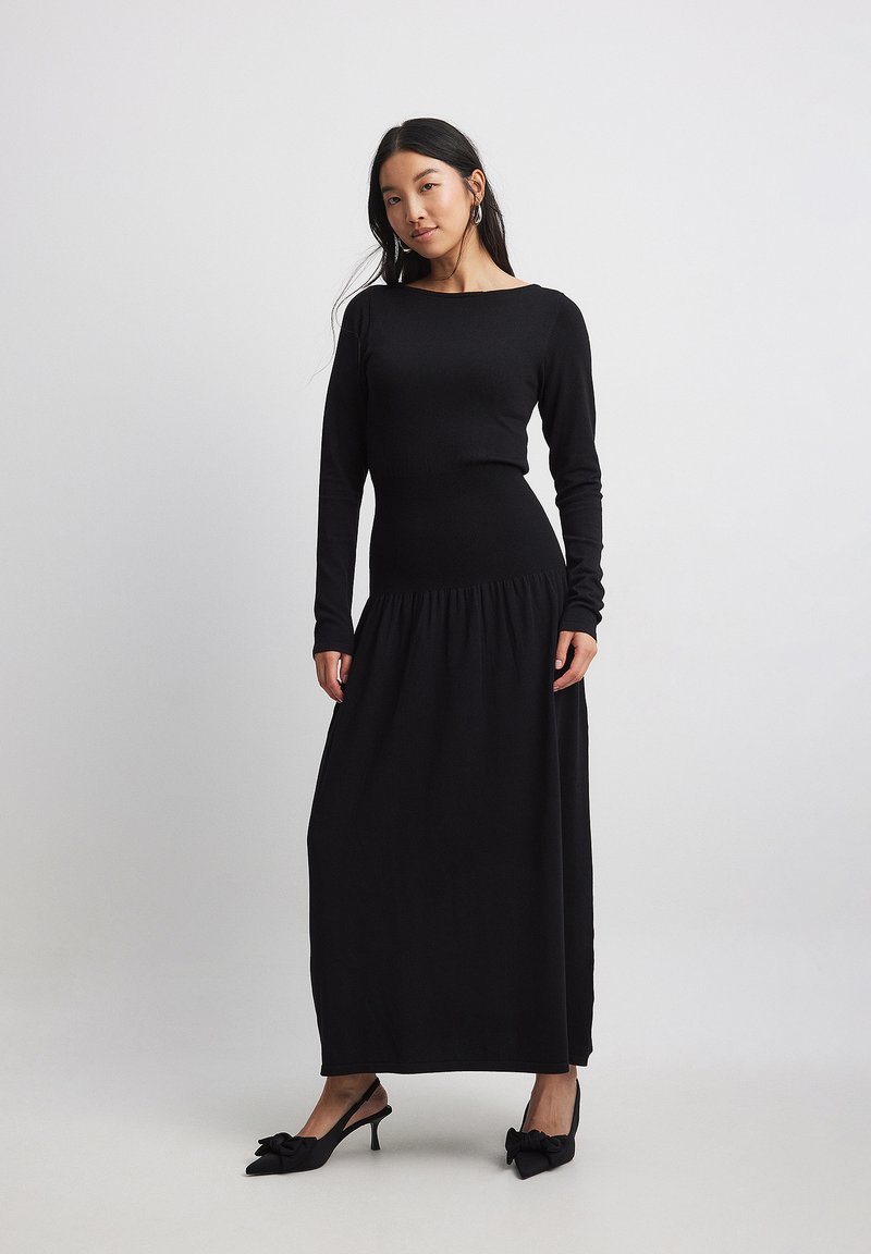 Black long-sleeve dress with a fitted top and gathered waist, flowing down to a straight skirt. Smooth fabric with minimal detailing.