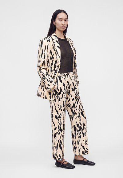 Femme aux longs cheveux noirs portant un costume beige et noir à motif abstrait sur un top noir transparent et des chaussures plates noires, debout les mains dans les poches.