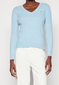 Osoba ubrana w jasnoniebieski sweter warkoczowy z dekoltem w serek i białe spodnie, stojąca na tle jasnego, jednolitego tła.