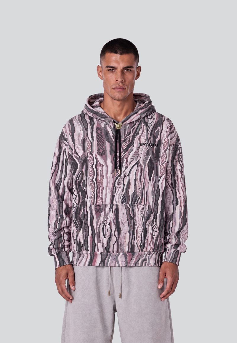 Homme aux cheveux courts portant un sweat à capuche à motifs rose, noir et gris et un pantalon gris clair, debout devant un fond uni.