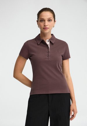 Mujer con una camisa polo marrón de manga corta con botones y pantalones negros, de pie frente a un fondo claro y liso.