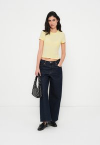 Keltainen istuva t-paita, jonka kanssa on tummansiniset leveälahkeiset farkut. Musta pilkullinen käsilaukku ja mustat kengät täydentävät asun. Valkoinen tausta.