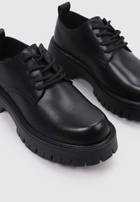 Zapatos de cuero negro con una superficie lisa, suela de goma con taco y cierre tradicional con cordones; presenta una punta redondeada y una lengüeta para tirar.