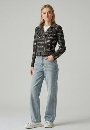 Frau steht mit der Hand in der Tasche, trägt eine schwarze, mit Nieten besetzte Lederjacke, hellblaue Jeans, weißes Hemd und weiße Turnschuhe vor einfarbigem Hintergrund.