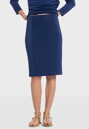 Tmavomodrá ceruzková sukňa z hladkej, elastickej tkaniny. Má pás s záhybmi a jemný akcent s logom. Kombinovaná so zlatými sandálmi na remienky.