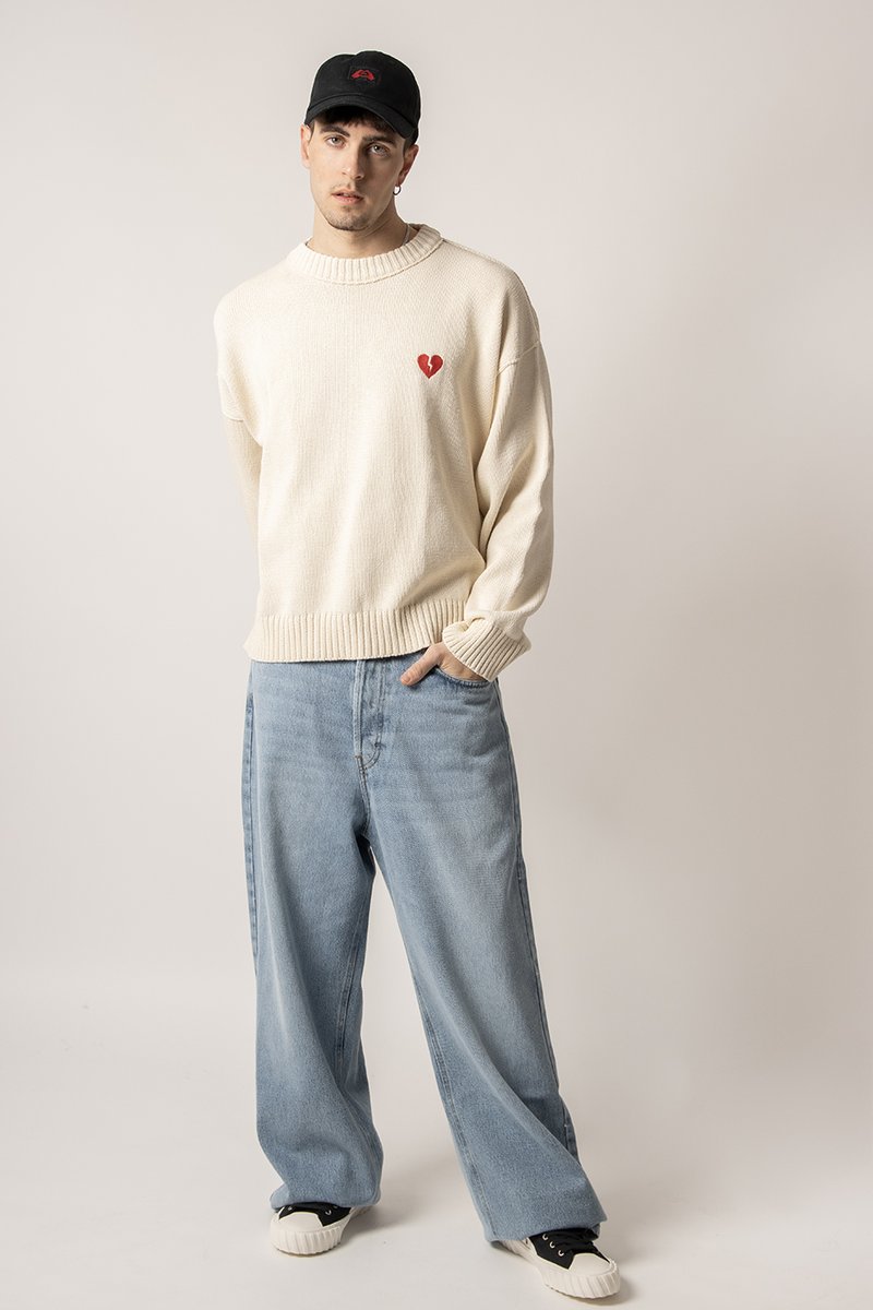 Chunky cream knit sweater with a red heart patch, paired with light blue wide-leg jeans and black cap, featuring a simple logo.