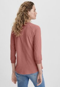 Camicetta rosa chiaro a maniche lunghe con maniche raglan, realizzata in tessuto morbido. La vista posteriore mostra un orlo arrotondato e semplici dettagli di cucitura.