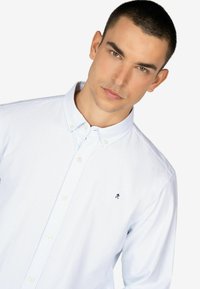 Camisa de botones de rayas azul claro con cuello, con botones blancos y un pequeño logo en el pecho. Textura de tela suave.