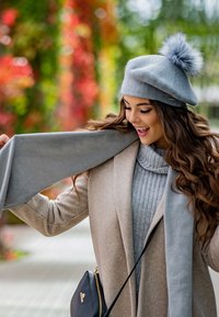 Beret en laine grise avec un pompon, écharpe grise et manteau beige clair sur un pull texturé. L'arrière-plan présente un feuillage coloré flou.