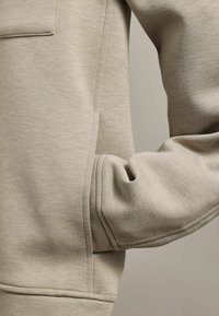 Tricou bej deschis cu o textură moale, având o mică buzunar frontal și o croială lejeră, cu cusături bine finisate și manșete ribbate.