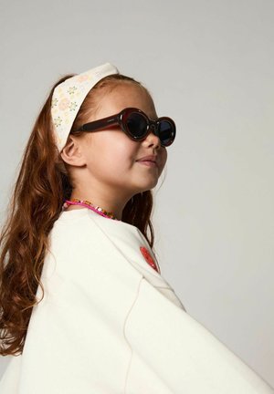 Ronde, donkere zonnebril met dikke, bruine monturen. Een crèmekleurige hoofdband met geborduurde bloemen. Het kind draagt een licht crèmekleurige top met een rood accent.