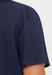 Navy blue blouse with puffed short sleeves, textured fabric, and a rounded neckline, showing subtle pleating at the shoulder seams.