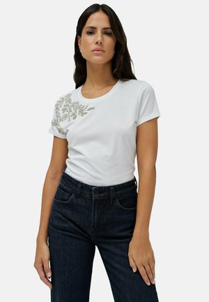 Camiseta de algodón blanca con mangas cortas y escote redondo, con adornos de cuentas plateadas en el hombro izquierdo y en la zona del pecho.