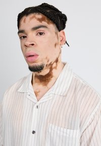 Skjorten har et lett, stripete design med knapper og en lomme på brystet. Modellen har flettet hår og synlig hudtonevariasjon i ansiktet og på nakken.