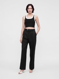 Νεαρή γυναίκα που στέκεται φορώντας μαύρο crop top χωρίς μανίκια με λευκή λεπτομέρεια, μαύρο παντελόνι ψηλόμεσο και λευκά ανοιχτά πέδιλα σε γκρι φόντο.