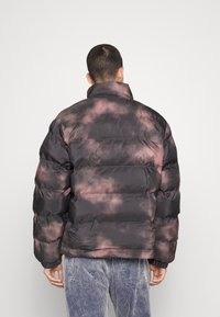 Hombre vistiendo una chaqueta acolchada de teñido anudado en negro y rosa vista desde atrás, combinada con pantalones de pana azul claro.