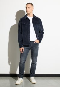 Mężczyzna w granatowej kurtce z materiału sztruksowego, białym T-shircie, wyblakłych czarnych dżinsach i białych trampkach stoi przy białej ścianie z cieniem.