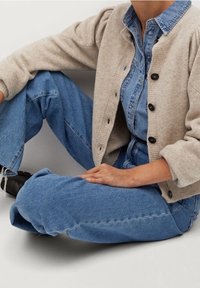 Cardigan en tricot beige avec col rond et boutons noirs superposé sur une chemise en denim bleu clair. Jean bleu à coupe décontractée.