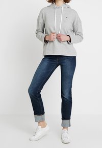 Felpa grigia chiara con tasca a canguro frontale, due cinturini bianchi e un piccolo logo. Abbinata a jeans in denim blu scuro e sneakers bianche.