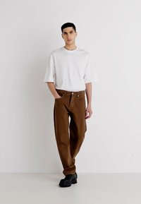 Bărbat îmbrăcat cu un tricou alb supradimensionat, pantaloni maro și pantofi negri, stând în fața unui perete alb simplu cu o mână în buzunar.