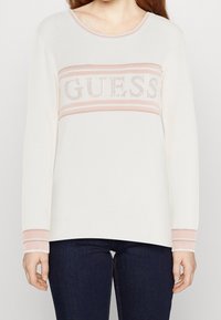 Pull blanc à manches longues avec accents roses. Logo "GUESS" en strass sur la poitrine. Poignets et encolure côtelés.