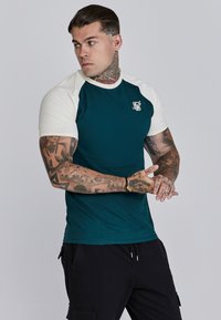 Donkergroene en crèmekleurige T-shirt met korte mouwen, voorzien van een getailleerd ontwerp en geborduurd logo op de borst. Zachte textuur en contrasterende mouwen.