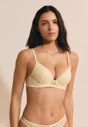 Młoda kobieta z długimi ciemnymi włosami, ubrana w jasnożółty koronkowy biustonosz i dopasowaną bieliznę na jasnobeżowym tle.