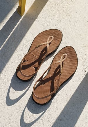 Par brun flad sandal med tynde remme, der kaster skygger på tekstureret lyst underlag nær gul stolben.