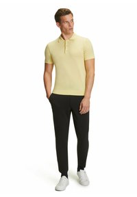 Maglietta polo gialla a maniche corte con colletto, abbinata a pantaloni neri a gamba affusolata e sneakers bianche. Caratterizzata da una vestibilità slim e una texture liscia.