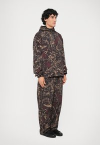 Set di due pezzi composto da una felpa con cappuccio e zip e pantaloni larghi. Il tessuto è scuro con motivi astratti in marrone e rosso. Scarpe nere.