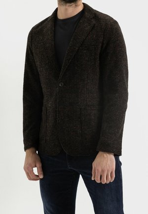Man wearing a dark textured blazer over a black shirt and dark blue jeans, standing against a plain light background.