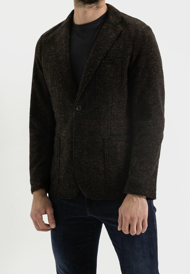 Homme portant un blazer texturé foncé sur une chemise noire et un jean bleu foncé, debout devant un fond clair uni.