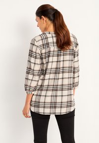 Beige geruite blouse met zwarte en bruine lijnen, driekwart mouwen en een afgeronde zoom. Getextureerde stof met een relaxte pasvorm. Zwarte broek.